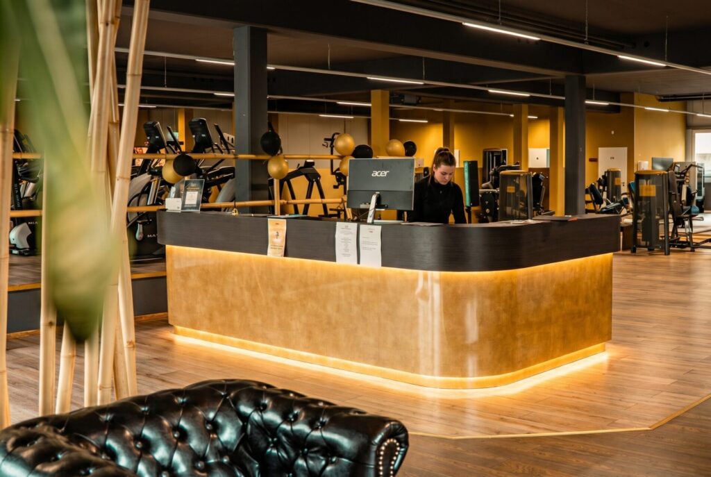 Empfangstresen im Limitless Fitnessstudio mit goldfarbenem Design, Chesterfield-Sofa und modernen Trainingsgeräten im Hintergrund